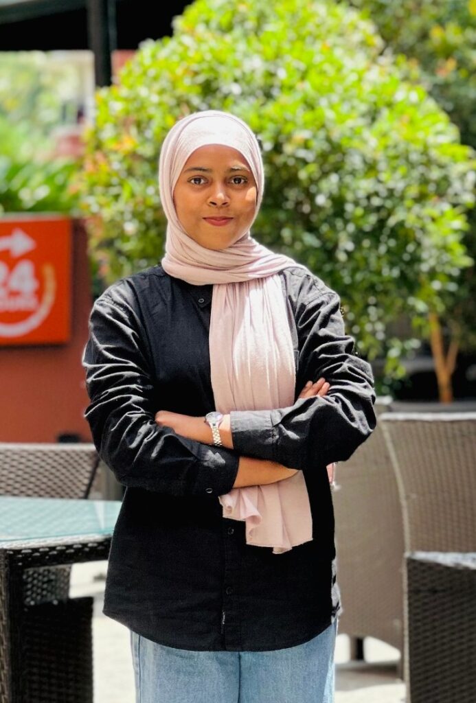 A confident young woman, known as the best digital marketing expert in Calicut, Kerala, stands outdoors in a modern café setting. She wears a light pink hijab, a black shirt, and blue jeans, embodying professionalism and expertise in digital marketing within the vibrant Calicut business scene